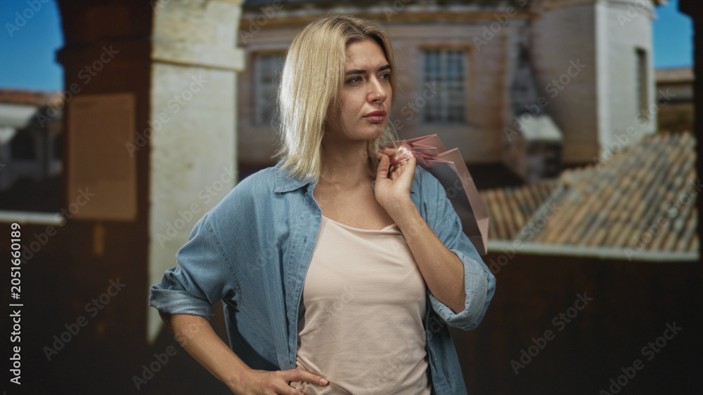 Fototapeta premium Woman holding pink shopping bag on shoulder with hand on hip in studio set with a projected building backdrop; pensive reflection.