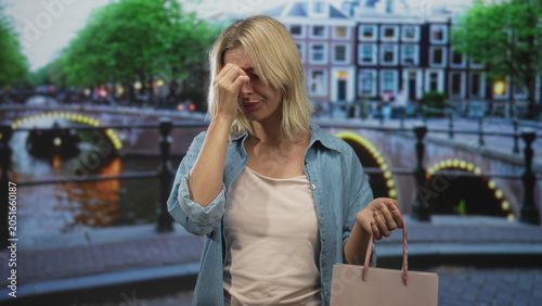 Foto Young blonde woman holding shopping bag, pinching bridge of nose with hand to face in street by canal and bridge lights; frustration errand