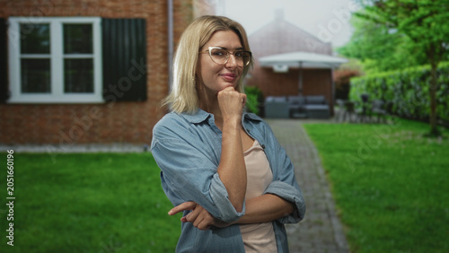 Photos Young blonde woman in glasses smiling with hand on chin and crossed arms, standing on a garden path in front of a brick building with patio umbrella; thoughtful confidence