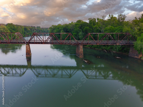 Rusty bridge spans calm, green river. Lush trees line both riverbanks. Reflections ripple gently below. Industrial decay meets natural beauty. Ideal for travel, nature, or nostalgia themes
