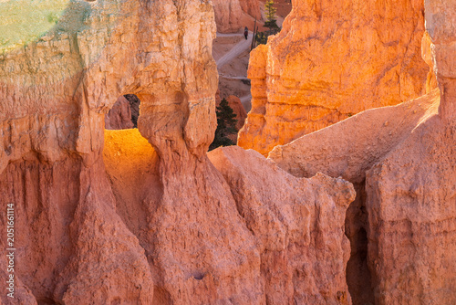 Golden light bathes towering red rock formations. Natural arches frame a narrow canyon path. Sunset hues cast warm tones across rugged sandstone. Texture details reveal erosion patterns over time