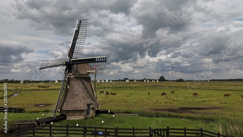 Billede på lærred Aerial view of the traditional Borgmolen windmill in Earnewald, surrounded by vibrant green meadows and wetlands under a dramatic cloudy sky, with distant sailboats