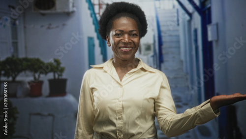 Obraz na płótnie Woman smiling and presenting an open right hand palm while wearing a beige shirt in a narrow white street with stairs and potted plants; friendly