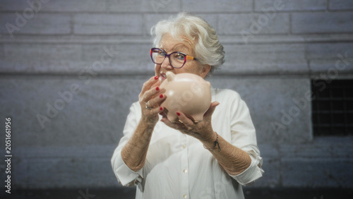 Принти на полотні Elderly woman holds piggy bank and gives thumbs up in front of building; savings independence optimism
