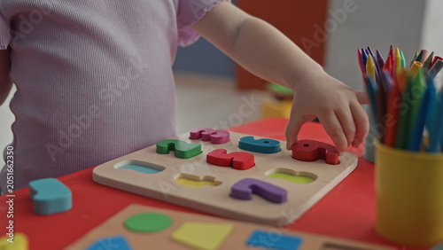 Toddler placing colorful wooden shape puzzle pieces with small hand on red play table in building; play learning concentration.