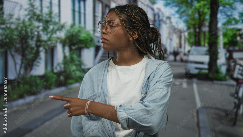 Fototapeta premium Woman with braided hair and glasses points finger to the right on street wearing denim shirt and white tee; hesitation concern.