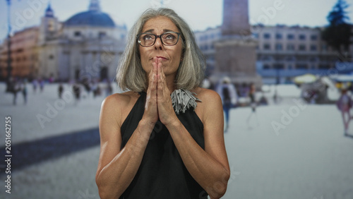 Tableau sur toile Middle age woman with grey hair and glasses, hands clasped in prayer in front of vatican building piazza, looking upward; faith devotion