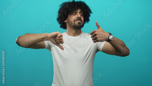 Slika na platnu Man showing both thumbs up and thumbs down gestures in studio with cyan backdrop; choice reflection ambivalence