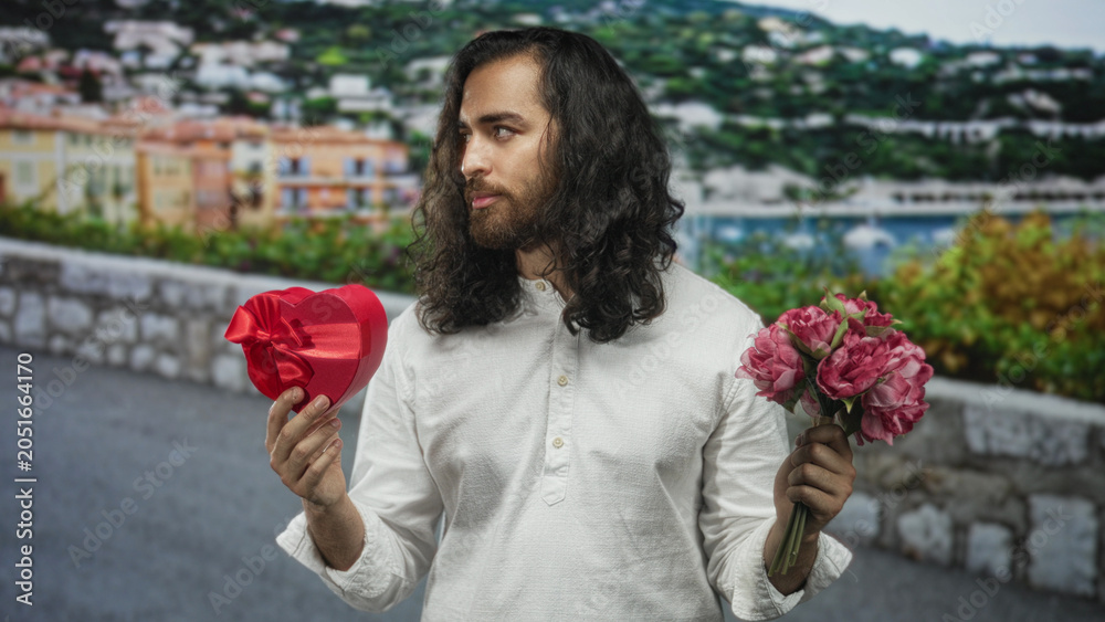 Fototapeta premium Man holding a heart shaped box and bouquet on a seaside street, holding gifts in both hands and glancing sideways; romance.