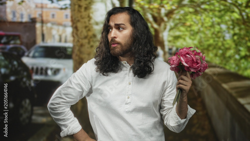 Konstfotografi Man with long hair and beard holds a pink bouquet in his hand and points finger toward the viewer on a street lane; pleading apology