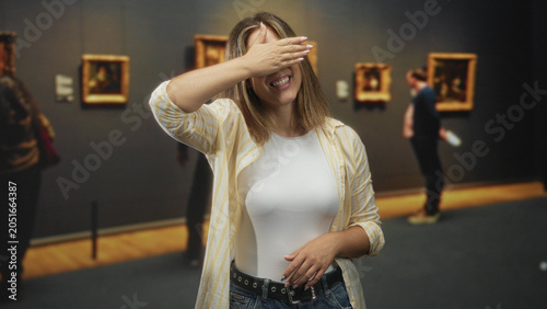 Billede på lærred Woman raising hand and briefly covering her eyes beside framed paintings in a museum gallery; playfulness curiosity
