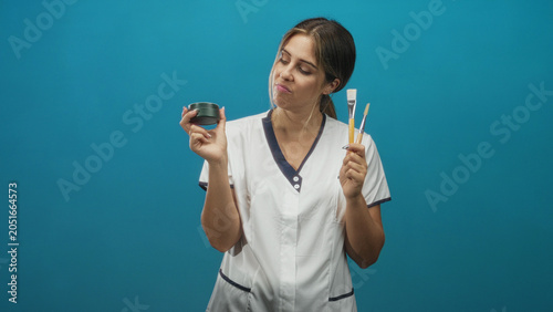 Foto Woman cosmetician in white tunic holds cream jar and three facial brushes, pouting and tilting head in studio; product doubt