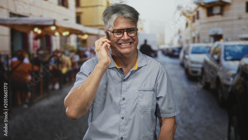 Konstfotografi Senior man holding smartphone to ear smiling while talking on a bustling street lined with parked cars and outdoor cafes; contentment connection
