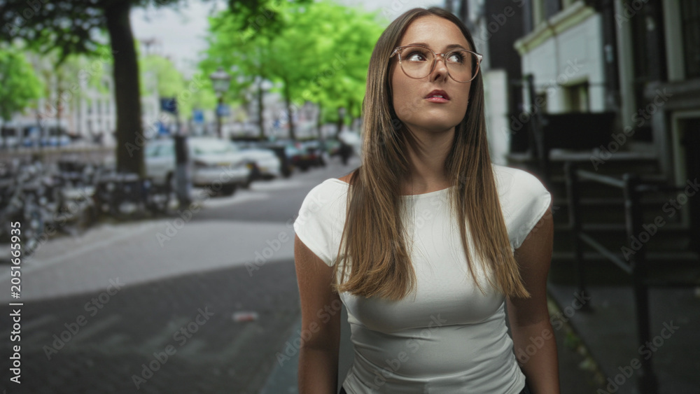 Fototapeta premium Young woman with glasses looking up while standing on a street beside parked bicycles and a building, wearing a white shirt and long hair; contemplation.