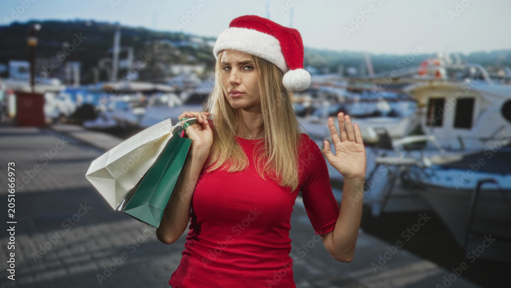 Fototapeta premium Woman holding shopping bags waving hand on street wearing santa hat with marina boats in view; holiday cheer.