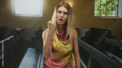 Foto Woman shaking fist and shouting, mouth open, standing among church pews by a stained glass window, wearing yellow pink sleeveless top; anger defiance