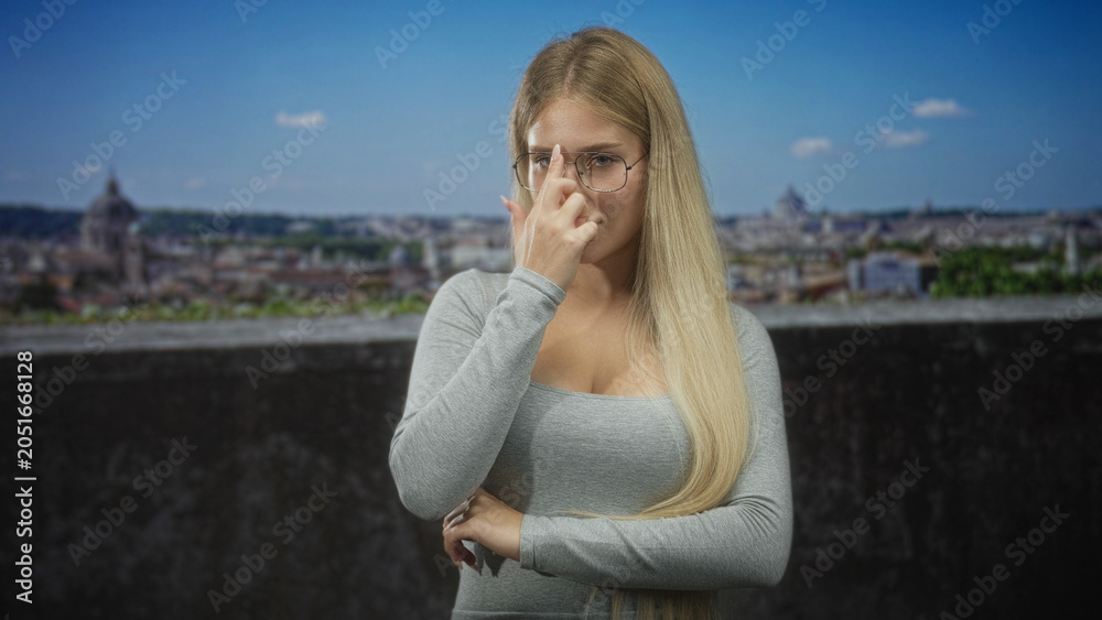 Obraz premium Woman touching glasses with finger at nose bridge on building terrace overlooking city; thoughtful contemplation.