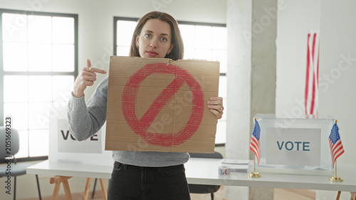 Bild auf Leinwand Woman young caucasian holding cardboard sign with red prohibition circle and points finger to sign in a voting building with ballot box and usa flags visible; defiance distrust