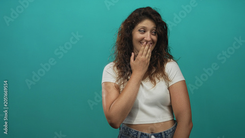 Quadro em tela Woman covering mouth with hand and smiling in teal studio, wearing white crop top and denim jeans; playful shyness