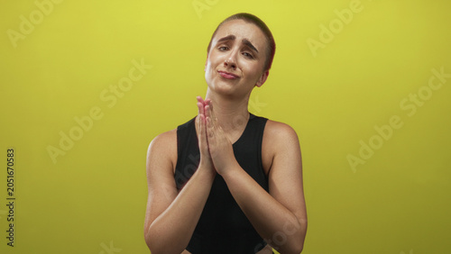 Konstfotografi Woman with short hair pressing palms together in a pleading prayer gesture, hands clasped in a studio with yellow backdrop; pleading
