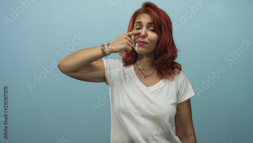 Fotografie Young hispanic woman pinching her nose and squinting while wearing a white t shirt in a light blue studio; disgust