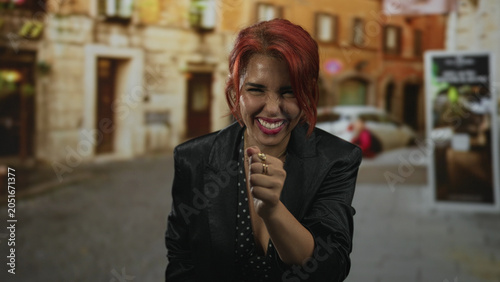 Konstfotografi Woman with red hair and hand to mouth laughing on a cobblestone street in front of buildings wearing blazer and rings; joy playfulness