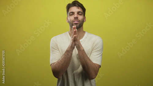 Tableau sur toile Man with beard presses hands together at mouth in studio against yellow backdrop, eyes closed in pleading gesture; quiet contemplation