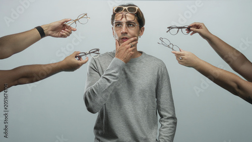 Bild auf Leinwand Man surrounded by hands offering eyeglasses, finger to chin gesture in studio; contemplation choice indecision