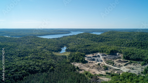Foto An expansive high-angle drone photograph showing an industrial open-pit gravel quarry and sand excavation site surrounded by dense rolling green hills and a large lake on the distant horizon