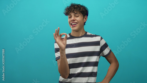Bild auf Leinwand Teenage boy makes ok hand sign with right hand near smiling face, wearing black and white striped tee, left hand on hip in turquoise studio; playful joy
