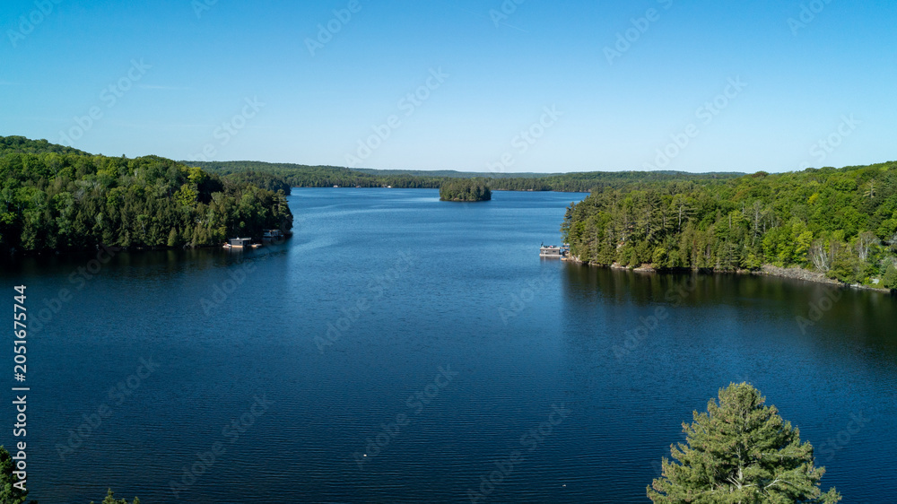 Fototapeta premium High-angle shot capturing a small, tree-covered island sitting in the middle of a deep blue lake, surrounded by expansive shorelines with cottages, docks and thick forest during a sunny summer day.