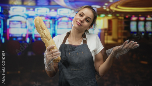 Bilde på lerret Woman baker in denim apron holds long baguette in one gloved hand and shrugs with visible face and slight smile beside row of slot machines in casino building; quirky bemused