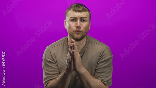 Foto Man young overweight pressing palms together in a prayer pose in a purple studio setting with visible beard and folded hands; pleading remorse vulnerability