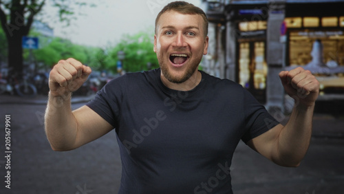 Εκτύπωση καμβά Man pumping fists and smiling on a city street with storefronts and bicycles in view; victory celebration