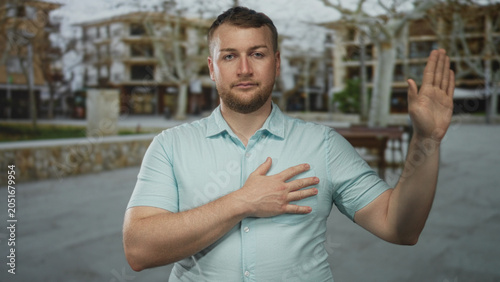 Εκτύπωση καμβά Young caucasian overweight man with hand on chest and raised palm standing in a street plaza in front of buildings and bare trees; respect sincerity