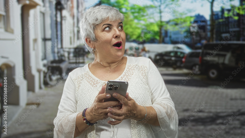 Fototapeta premium Senior woman holding smartphone, hands tapping screen on a city street while smiling and reading messages from phone; surprise connection.