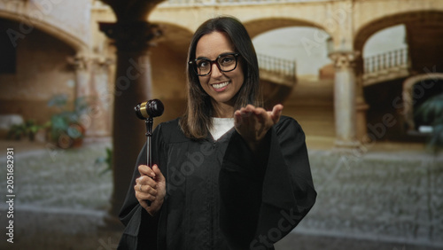 Fotografie Woman judge in black robe holds gavel and offers open hand gesture toward viewer in a historic court building, smiling with glasses; confidence