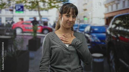 Obraz na płótnie Woman hand on chest gesture in street with parked cars, planters and building facades; gratitude sincerity