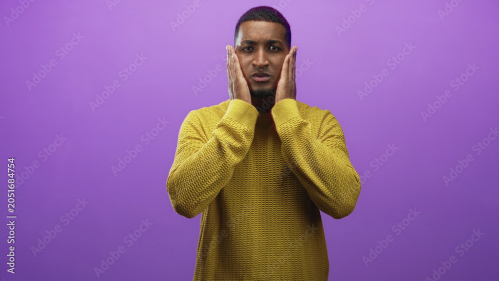 Obraz premium Man pressing hands to face in purple studio wearing yellow sweater and showing tense expression; surprise.