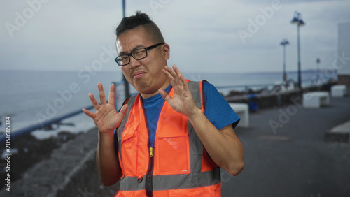 Tablou pe pânză Man in orange reflective vest clasping hands near chest on a street promenade by the seaside with tense face and grimace; unease