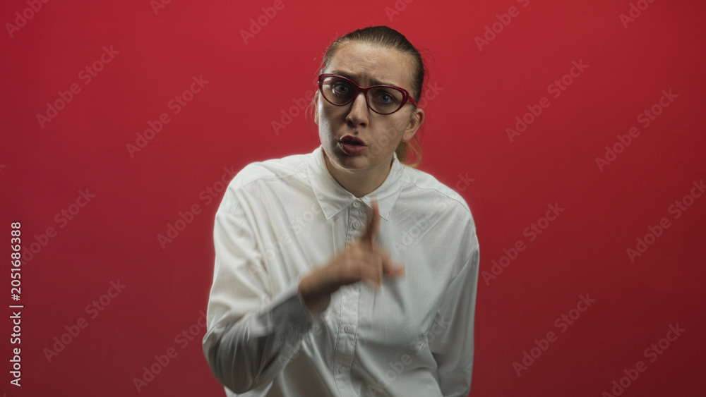 Obraz premium Woman with red glasses pointing finger and speaking, white button shirt, neutral puzzled expression against a red studio wall; uncertainty inquiry.