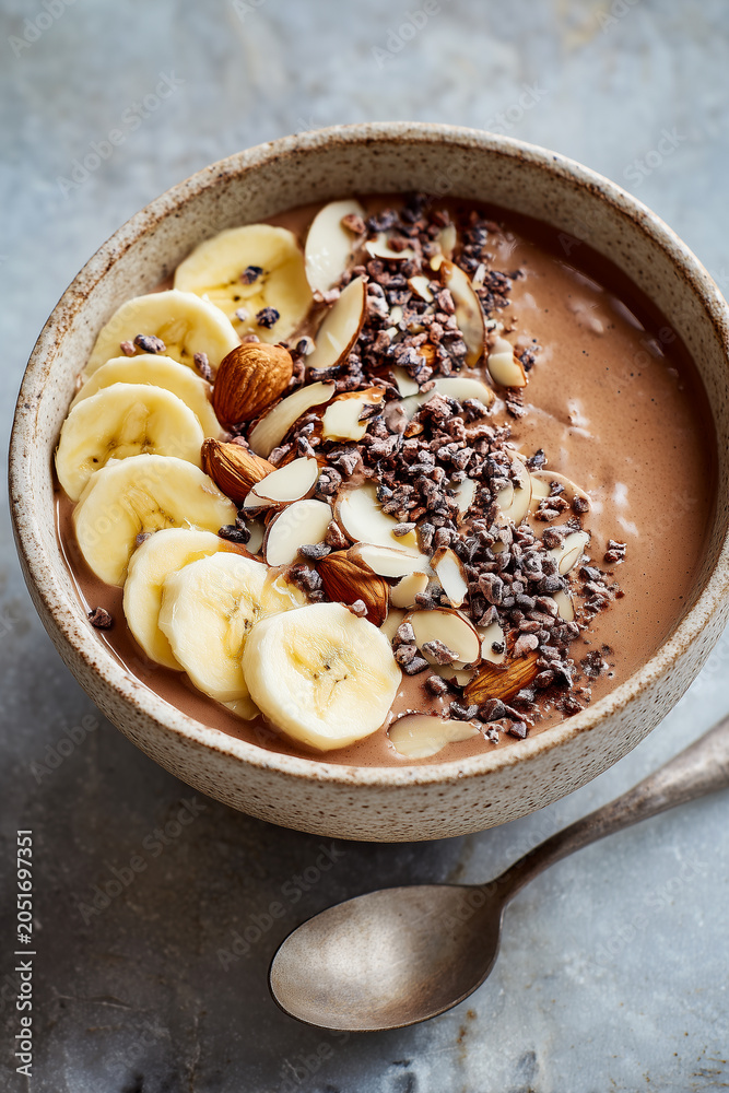 Fototapeta premium Healthy chocolate smoothie bowl topped with fresh sliced bananas, almonds, and cacao nibs in a ceramic bowl. Perfect delicious and nutritious breakfast