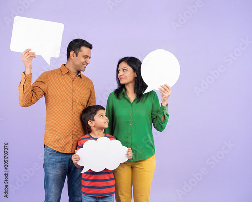 Happy Hispanic family holding empty white speech bubble signs on lilac background. Father, mother and little son smiling together with communication, horizontal background with place for text.