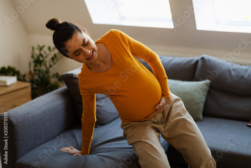 Foto Pregnant woman straining to stand up from sofa, hand pressed to abdomen