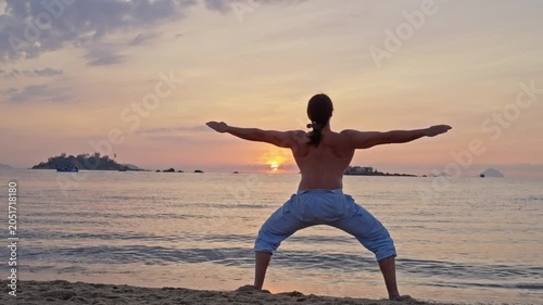 Silhouette stretching on a beach at sunset. Back view of a silhouetted person standing on the sandy shore with arms extended toward a calm sea, glowing sunset, distant islands, and a peaceful horizon
