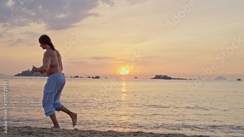 Shirtless man running along a beach at sunset. A shirtless adult man runs along a sandy beach at sunset, with calm water, distant islands, and a warm golden sky creating a peaceful coastal scene