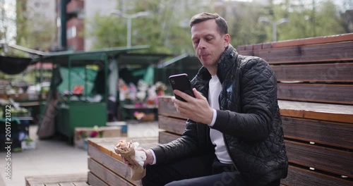 A man sits on wooden seating in an urban park, enjoying a snack while focused on his smartphone. The environment is vibrant, with market stalls and greenery nearby