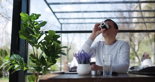 A man is focused on his work, sitting at a table in a cafe surrounded by greenery. Bright sunlight filters through the glass structure, creating a warm atmosphere for productivity and relaxation