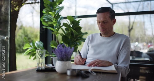 A man sits at a wooden table in a cafe, writing in a notebook while enjoying the company of indoor plants. Sunlight streams in, creating a warm atmosphere