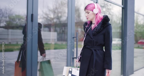 Model with pink hair walks casually outside a modern building wearing headphones while carrying stylish bags in a chic outfit